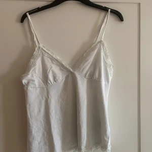 Vit spetskantad linnetopp - Säljer en vit linnetopp med tunna axelband och v-ringning. Toppen har fina spetsdetaljer.