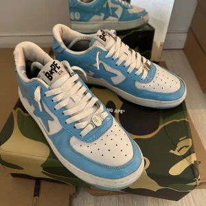 Bape Sta Baby Blue  - Säljer ett par Bape Sta sneakers i blått och vitt med klassisk stjärndesign på sidan. Skorna har snörning, perforerad tå och platt sula. Ovandelen är i glansigt syntetmaterial och innerfodret är vitt. Perfekta för dig som gillar streetwear och unika sneakers.