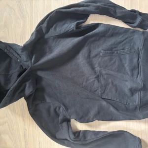 Svart hoodie med ficka framtill - En svart hoodie med klassisk känguruficka framtill och huva. Tröjan har långa ärmar och ribbade muddar vid ärmslut och nederkant. Perfekt för chill dagar eller när du vill ha något bekvämt och enkelt.