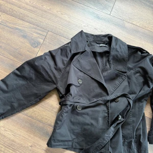 Svart trenchcoat med bälte - Stilren svart trenchcoat med klassisk krage, dubbelknäppning och tillhörande bälte i midjan. Jackan har långa ärmar och knappar framtill.