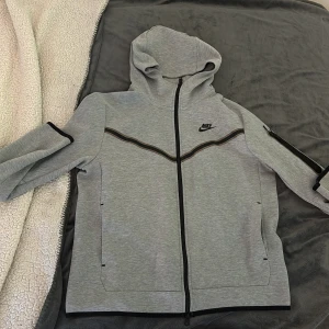 Grå Nike hoodie med dragkedja M - Snygg grå hoodie från Nike i storlek M med hel dragkedja och svart Nike-logga på bröstet. Tröjan har huva, svarta detaljer längs ärmar och bröst samt praktiska fickor. Perfekt för en sportig och avslappnad stil.