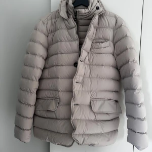 Beige pufferjacka från Herno - Säljer en beige pufferjacka från Herno med quiltad design och hög krage. Jackan har både dragkedja och knappar framtill, samt stora fickor med lock och en bröstficka. Perfekt för kyliga dagar och riktigt skön att bära. Är storlek 48 men lite mindre i storleken