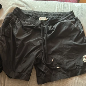 Svarta Moncler shorts med snörning, skön material bra att ha på sommaren. Använde den inte mycket och passar inte min storlek.  - Snygga svarta shorts från Moncler med elastisk midja och snörning framtill. Shortsen har Moncler-logga på benet och är tillverkade i ett glansigt syntetmaterial, perfekta för sommaren eller stranden. Klassisk passform och sportig vibe.