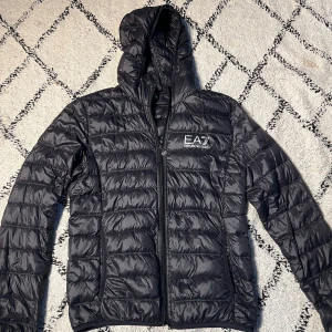 Svart pufferjacka från EA7 Emporio Armani - Snygg svart pufferjacka från EA7 Emporio Armani med huva och dragkedja framtill. Jackan har quiltad design och logga på bröstet. Perfekt för kyliga dagar och ger en sportig vibe. Materialet är glansigt och lätt, vilket gör den smidig att bära.
