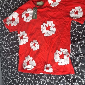 Röd blommig t-shirt från Denim Tears - Röd t-shirt från Denim Tears med stora vita blommor tryckta över hela plagget. Klassisk passform och rund hals. Perfekt statement-piece för dig som vill sticka ut med en färgstark och unik stil.