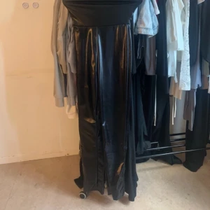 Svart Bodysuit - Säljer en svart Bodysuit  i glansigt syntetmaterial med hög midja och slits framtill. Kjolen har en cool, edgy look och passar perfekt om du vill sticka ut. Materialet är blankt och ger en riktigt snygg finish.