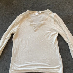 Vit långärmad blus med spets - Supersöt vit långärmad blus från KappAhl med v-ringning och bred spetskant framtill. Topp i mjukt material som känns skönt mot huden och har en feminin touch med den blommiga spetsen. Perfekt att styla med jeans eller kjol.