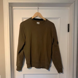 Mörkgrön sweatshirt från C.P. Company - Mörkgrön sweatshirt från C.P. Company med klassisk rund hals och lång ärm. Tröjan har en ikonisk svart linsdetalj på ärmen och en diskret ficka med dragkedja. Tillverkad i mjuk bomull för en skön och avslappnad stil. Hör av dig vid eventuella frågor eller funderingar! Pris är diskuterbart!