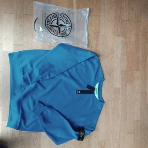 Blå sweatshirt från Stone Island - Blå sweatshirt från Stone Island med klassisk rund hals och ribbade muddar. Ikonisk logotyp-patch på vänster ärm. Tröjan är i mjuk bomull och har en clean, stilren look som passar perfekt till jeans eller joggers.