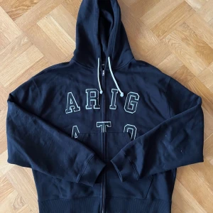 Svart hoodie med tryck från Axel Arigato - Svart AA Legendz hoodie med vit snörning i huvan och stort tryck 'ARIGATO' på bröstet. Tröjan har dragkedja framtill, känguruficka och ribbade muddar. Perfekt för en chill och avslappnad stil. Nypris 2500kr 