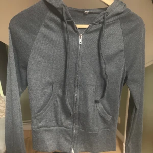 Mörkgrå hoodie med dragkedja - Mörkgrå hoodie med dragkedja framtill och huva med snören. Tröjan har två fickor på magen och är ribbad i materialet. Långärmad och i en croppad modell som ger en snygg och avslappnad look.
