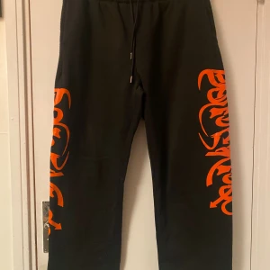 Svarta mjukisbyxor Manfinity XL - Svarta mjukisbyxor från Manfinity i storlek XL med feta orangea tryck längs båda benen. Byxorna har dragsko i midjan och är gjorda i mjukt material, perfekta för chill eller streetwear. Snygg kontrast mellan svart tyg och det orangea mönstret.