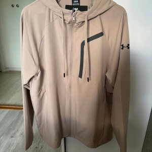 Under Armour jacka - Säljer en beige vindjacka från Under Armour med dragkedja och huva. Tröjan har långa ärmar, svarta detaljer på bröstet och logga på ärmen. Perfekt för träning eller chill dagar. Materialet är mjukt och lätt, troligen polyester.
