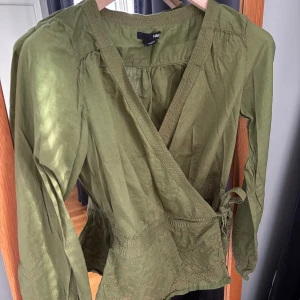 Olivgrön omlottblus från H&M - Olivgrön omlottblus från H&M med knyt i sidan och broderade detaljer nertill. Blusen har v-ringning, långa ärmar med resår vid ärmslut och smock i midjan för en snygg passform. Perfekt att styla med jeans eller kjol.