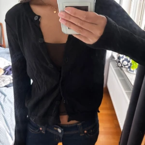 Svart kofta med knappar - En svart, klassisk kofta från Dagmar med rund hals och långa ärmar. Koftan har flera svarta knappar framtill och är tillverkad i ett tunt, stickat material. Perfekt att slänga över en topp eller linne för en enkel och stilren look.