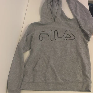 Grå hoodie från FILA med logga - Säljer en grå hoodie från FILA med stor svart logga på bröstet. Tröjan har huva med snörning och en stor magficka. Perfekt för dig som gillar sportig och avslappnad stil. Dock är snörena borta de syns på bilden🩷