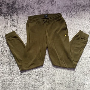 Olivgröna mjukisbyxor Lyle & Scott - Olivgröna mjukisbyxor från Lyle & Scott med klassisk gul logga broderad på benet. Byxorna har resår och snörning i midjan samt muddar vid bensluten. Tillverkade i mjukt bomullsmaterial för maximal komfort.