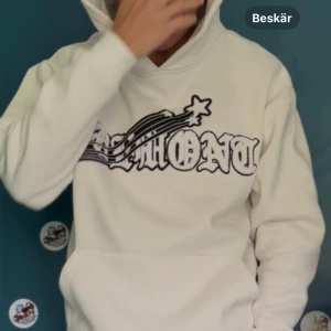 Demont tröja  - Vit hoodie med stor svartvit grafisk text och stjärnshooting-tryck över bröstet. Klassisk känguruficka framtill och huva. Oversized passform och mjukt material, perfekt för chill dagar.
