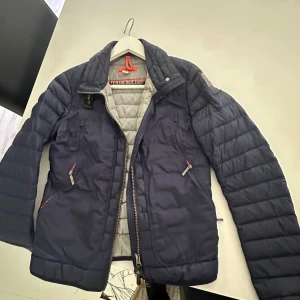 Parajumpers jacka - Säljer en mörkblå pufferjacka från Parajumpers i storlek S. Jackan har quiltad design, dragkedja framtill, flera fickor med dragkedja och en snygg patch på ärmen. Insidan är ljusgrå och jackan har orange detaljer. Perfekt för kyliga dagar! 
