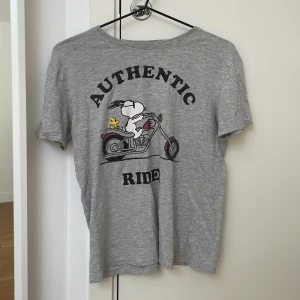 Grå Peanuts t-shirt från Zara - Grå pyamas T-shirt från Zara med tryck av Snoopy och Woodstock på motorcykel och texten 'AUTHENTIC RIDE'. Klassisk rund hals och korta ärmar. Materialet är mjuk bomull, perfekt för en chill och avslappnad stil.