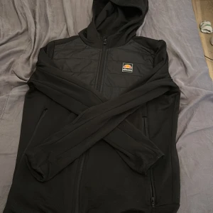 Svart hoodie från Ellesse med quiltad front - Svart hoodie från Ellesse med quiltad front och ryggparti. Tröjan har dragkedja framtill, huva och två sidofickor med dragkedja. Perfekt för kyliga dagar och har en sportig vibe med Ellesse-logga på bröstet. Det är största barnstorlek men det är extra Small.