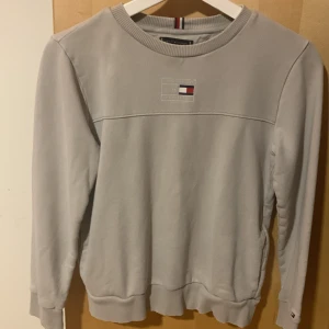 Ljusgrå sweatshirt från Tommy Hilfiger - Snygg ljusgrå sweatshirt från Tommy Hilfiger med broderad logga på bröstet och klassiska ribbade muddar. Tröjan har rund halsringning och diskreta Hilfiger-detaljer i nacken. Perfekt för en chill och stilren look.