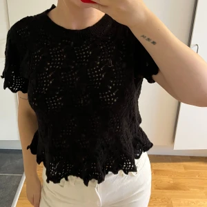 Svart virkad topp med snörning bak - Svart kortärmad virkad topp med vågig kant och snygg snörning i ryggen. Toppen har ett genombrutet mönster och är croppad med rund halsringning. 