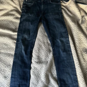 Mörkblå jeans från Nudie  - Säljer ett par mörkblå jeans från Nudie Jeans i klassisk femficksmodell. Jeansen har en slim passform och raka ben, med snygga slitningar och detaljerade sömmar. Perfekta för dig som gillar stilrena och tidlösa jeans. Storlek 27/28. Storleken är mer som 28 30  men tajta 