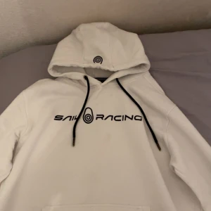 Vit hoodie från Sail Racing - Säljer en clean vit hoodie från Sail Racing med svart logga och tryck på bröstet. Tröjan har huva med svarta snören och en stor magficka. Perfekt för dig som gillar en sportig och stilren look. Funkar till L