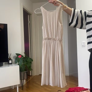 Ljus långklänning med plissering - Säljer en elegant ljus långklänning med plisserad överdel och markerad midja med fina rynkdetaljer. Klänningen är ärmlös och har ett mjukt, flytande tyg som ger en romantisk känsla. Perfekt för speciella tillfällen eller när du vill känna dig extra fin.