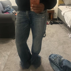 Baggy blå jeans från Ninth - Baggy blå jeans från Ninth med slitna detaljer och flera fickor på sidorna. Jeansen har låg midja och breda ben med fransad nederkant. Perfekta för en avslappnad streetstil och passar dig som gillar loose fit och vintagevibbar.