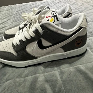 Nike Dunk sneakers i grått och vitt - Nike Dunk sneakers i grå mocka och vitt skinn med svart swoosh och cool björndetalj på hälen. Skorna har vita snören, perforerad tå och färgglada detaljer på plösen. Perfekta för dig som gillar streetstyle och unika detaljer.