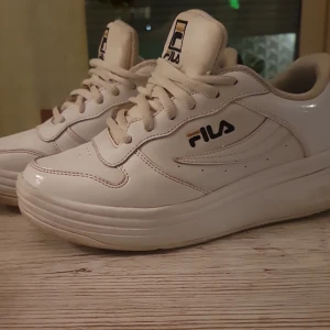 Vita Fila sneakers - Säljer ett par klassiska vita Fila sneakers med snörning och rund tå. Skorna har Fila-logga både fram och bak samt detaljer i svart och guld. Ovandelen är i syntetmaterial och de har en platt sula. Perfekta för en clean och sportig stil.
