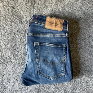 Två par jeans från Jack & Jones - Säljer två par snygga jeans från Jack & Jones. Ett par är klassiskt blå med kontrastsömmar och läderpatch, det andra paret är mörkgrått med svart patch. Båda har femficksdesign och raka ben, perfekta för en stilren look.