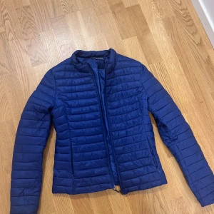 Blå pufferjacka från Blue Sand - Säljer en stilren blå pufferjacka från Blue Sand. Jackan har horisontella sömmar, hög krage och dragkedja framtill. Perfekt för kyliga dagar och enkel att matcha med olika outfits. Lätt och smidig att bära med sig. Storlek S XS.