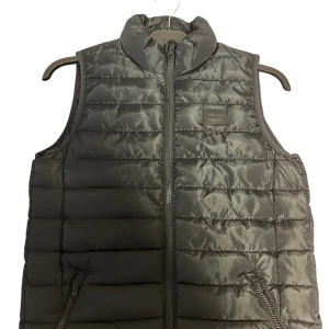 Svart dunväst från Jack & Jones - Snygg svart dunväst från Jack & Jones med quiltad design och hög krage. Västen har dragkedja framtill och två fickor med dragkedja. Perfekt att slänga över en hoodie för en cool look. Materialet är glansigt och känns lätt men ändå värmande. Till pojkar storlek 152