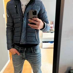 Blå quiltad pufferjacka Superdry - Säljer en blå quiltad pufferjacka från Superdry, modell Fuji. Jackan har ett diskret mönster, dragkedja och knappar framtill samt två fickor med dragkedja. Perfekt för kyligare dagar och har en snygg, sportig look.