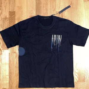 Svart AMIRI t-shirt med tryck - Svart t-shirt från AMIRI med stort tryck på ryggen och mindre tryck på bröstet. Trycket har en cool drip-effekt i vitt, blått och rött. Klassisk rund hals och korta ärmar. Perfekt för dig som gillar streetwear och vill sticka ut.