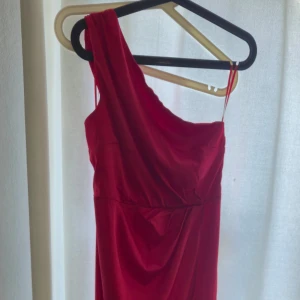 Röd one shoulder klänning från Nelly - Snygg röd klänning från Nelly i one shoulder-modell med draperad detalj framtill. Klänningen har en tight passform och är tillverkad i ett mjukt, slätt material som känns lyxigt. Perfekt för dig som vill sticka ut med en trendig och elegant look.