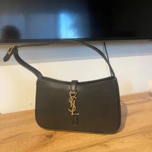 Svart axelväska  - Snygg svart axelväska i slätt skinn med ikoniskt YSL-spänne i guld framtill. Väskan har en stilren, rektangulär form och justerbart axelband. Invändigt finns en dragkedjeficka och diskret Saint Laurent Paris-logga.
