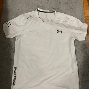 Vit Under Armour tränings t-shirt  - Hej! Jag Säljer nu min vita Under Armour t-shirt i funktionsmaterial, perfekt för löpning eller gym. T-shirten har korta ärmar, rund hals och reflekterande loggor på bröstet, axeln och längs sidan. Snygg och sportig design som andas och transporterar bort fukt. Vid frågor så är det bara att ställa! Tröjan sitter som slim fit! Tags medföljer.
