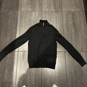 Mörkgrå stickad half zip-tröja - En mörkgrå Morris stickad tröja med half zip och hög krage. Tröjan har lång ärm och ribbade muddar vid ärmslut och nederkant. Perfekt för lager-på-lager och snygg till både jeans och chinos.