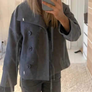Grå kort kappa från Gina Tricot - Säljer en snygg grå kort kappa från Gina Tricot med bred krage och dubbelknäppning framtill. Jackan har raka ärmar och en stilren, boxig passform som ger en cool vibe. Perfekt för dig som gillar minimalistisk stil och vill ha något som sticker ut lite extra. Kontakta för fler bilder och frågor🤍