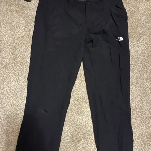 Svarta täckbyxor från The North Face - Svarta täckbyxor från The North Face med klassisk logga på benet. Byxorna har raka ben, knapp och dragkedja i midjan samt praktiska fickor. Perfekta för kalla dagar och outdoor-äventyr.  Så som det syns på bilden har det skett en lite slit skada därav priset