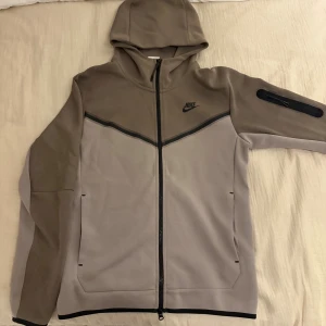 Nike tech i beige och grå, stl S - Nike hoodie i beige och grå med svart logga på bröstet. Tröjan har hel dragkedja, huva och en praktisk ficka med dragkedja på ena ärmen. Perfekt för chill eller träning, snygg och sportig look. Tveka inte om ni har mera frågor!😄