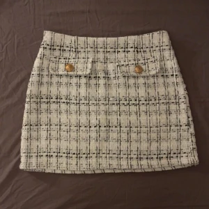 Rutig kortkjol från SHEIN i tweed - Superfin kortkjol från SHEIN i vit och svart rutig tweed. Kjolen har två dekorativa fickor framtill med guldknappar och stängs med en diskret dragkedja i sidan. Perfekt för en chic och trendig look.