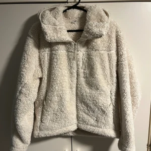 Vit teddyjacka från Uniqlo XS - Supermjuk och fluffig vit teddyjacka från Uniqlo i storlek XS. Jackan har hög krage, hel dragkedja framtill och praktiska fickor med dragkedja. Perfekt för kyliga dagar när du vill vara både varm och stylish.