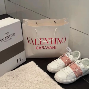 Valentino Rockstud sneakers vit/rosa - Snygga Valentino Garavani Rockstud sneakers i vitt skinn med ljusrosa band och ikoniska nitar. Klassisk rund tå, platt sula och vita snören. Perfekta för dig som vill ha en lyxig och trendig look med extra edge.