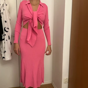 Rosa ribbad långklänning med knyt - Säljer en superfin rosa långklänning i ribbad bomullsmix med lång ärm, krage och snygg knytning framtill. Klänningen har cut out-detaljer vid midjan och sitter figurnära för en riktigt trendig look.