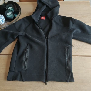 Svart hoodie från Nike med dragkedja - Säljer en fet Nike Tech den här två super små skador som inte syns när man använder den. Man får med en gåva om man köper